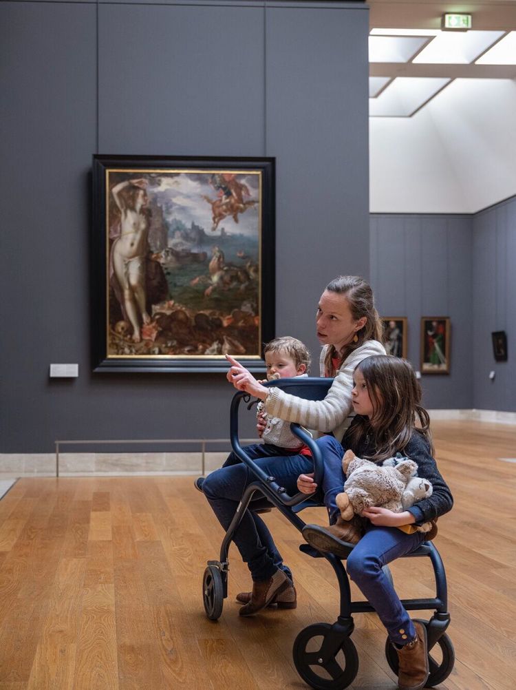 La foto riprende una giovane donna seduta con in braccio un bambino di circa 2 anni su una specie di sedia mobile con le ruote (una delle Flauneuses del Louvre) che le permette di sedersi davanti alle opere del museo. Accanto a lei, su un'altro braccio della stessa sedia, è seduta una bambina di circa 5 anni che guarda verso l'opera d'arte indicata dalla madre