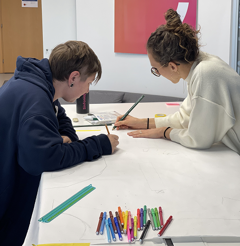 La foto mostra un momento di preparazione di un workshop di service design. Due ragazze stanno disegnando su un grande foglio bianco con pennarelli e matite