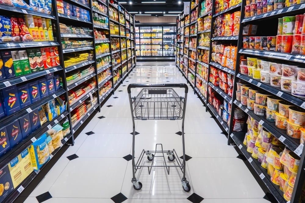 Foto di un carrello in mezzo ad una corsia di un supermercato, gli scaffali ai lati sono pieni di prodotti